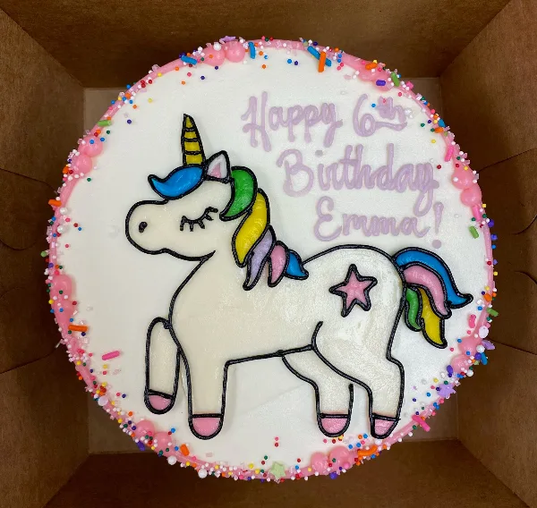 A round white buttercream cake featuring a piped unicorn design, colorful sprinkles, and the text 'Happy 6th Birthday Emma!'