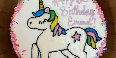A round white buttercream cake featuring a piped unicorn design, colorful sprinkles, and the text 'Happy 6th Birthday Emma!'