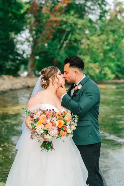 A bride in a white gown and a groom in a green suit kissing outdoors near a body of water.