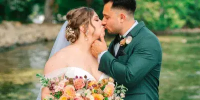 A bride in a white gown and a groom in a green suit kissing outdoors near a body of water.
