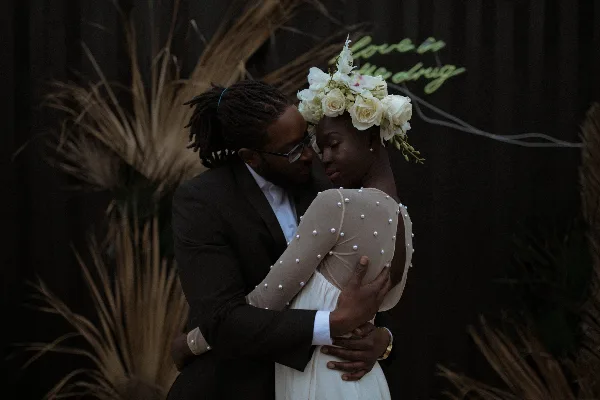A groom in a dark suit and a bride in a pearl-embellished dress embrace in front of a dark floral backdrop.