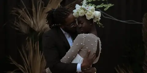 A groom in a dark suit and a bride in a pearl-embellished dress embrace in front of a dark floral backdrop.