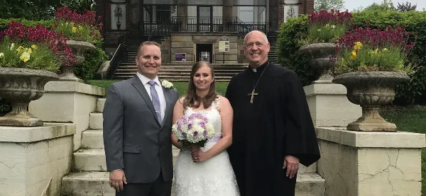 A wedding officiant in black clerical robes standing with a bride and groom in front of a stone building.