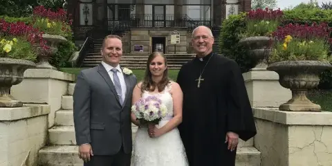 A wedding officiant in black clerical robes standing with a bride and groom in front of a stone building.