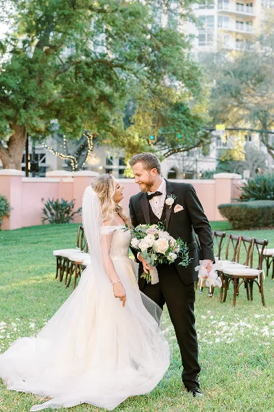A bride in a white gown and a groom in a black tuxedo standing together on a grassy lawn with wedding chairs in the background.