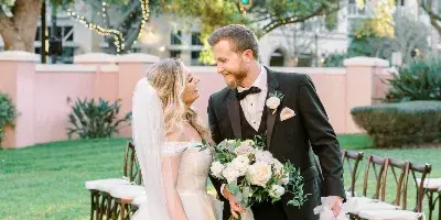 A bride in a white gown and a groom in a black tuxedo standing together on a grassy lawn with wedding chairs in the background.