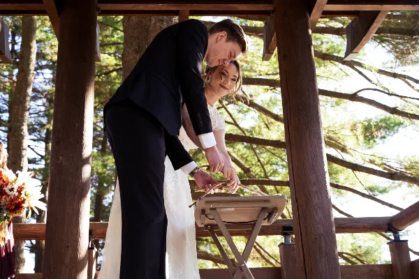 A bride and groom performing a unity candle ceremony at an outdoor wooden pavilion.
