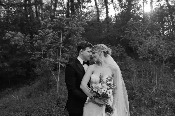 Black and white portrait of a bride and groom standing close together in a wooded area, with the bride holding a bouquet.
