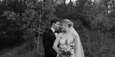Black and white portrait of a bride and groom standing close together in a wooded area, with the bride holding a bouquet.