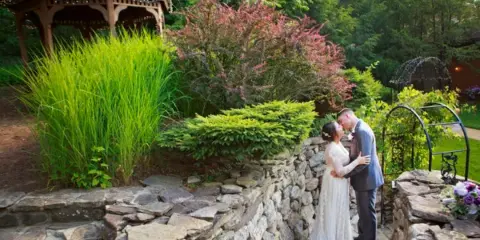 A bride and groom sharing a kiss on a stone staircase in a garden setting with a gazebo in the background.