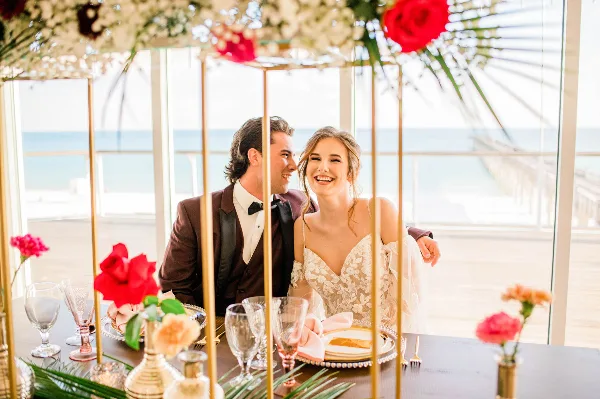 A bride and groom seated at a decorated reception table inside a glass-walled venue with a view of the ocean.