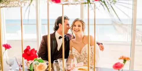 A bride and groom seated at a decorated reception table inside a glass-walled venue with a view of the ocean.