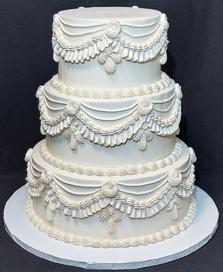 A 3-tier white wedding cake featuring elaborate buttercream piping, swags, and floral details.