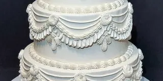 A 3-tier white wedding cake featuring elaborate buttercream piping, swags, and floral details.
