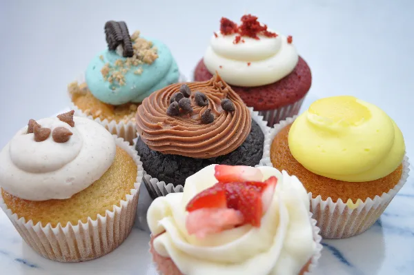 An assortment of six decorated cupcakes featuring different frosting colors and toppings, including chocolate chips, strawberries, and cookie pieces.