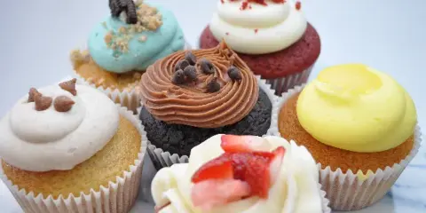 An assortment of six decorated cupcakes featuring different frosting colors and toppings, including chocolate chips, strawberries, and cookie pieces.