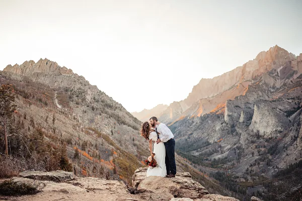 A bride and groom embracing on a rocky mountain overlook with a valley in the background.