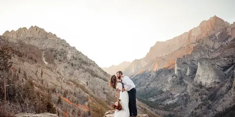 A bride and groom embracing on a rocky mountain overlook with a valley in the background.