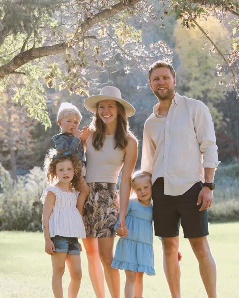 A family of five posing outdoors in a sunlit park setting.