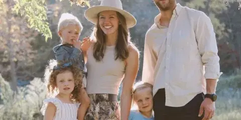 A family of five posing outdoors in a sunlit park setting.