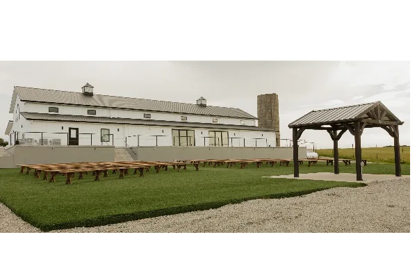 Exterior view of a white barn-style wedding venue with a wooden gazebo and ceremony seating on a lawn.