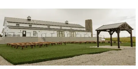 Exterior view of a white barn-style wedding venue with a wooden gazebo and ceremony seating on a lawn.