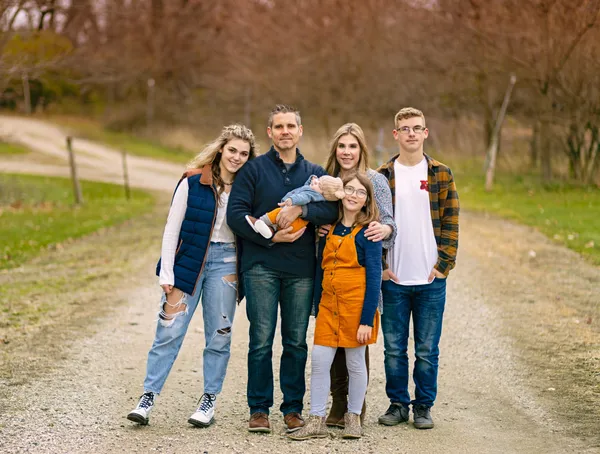 A family of six, including a man holding a baby, posing together on a dirt path outdoors.