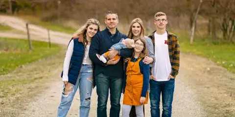 A family of six, including a man holding a baby, posing together on a dirt path outdoors.