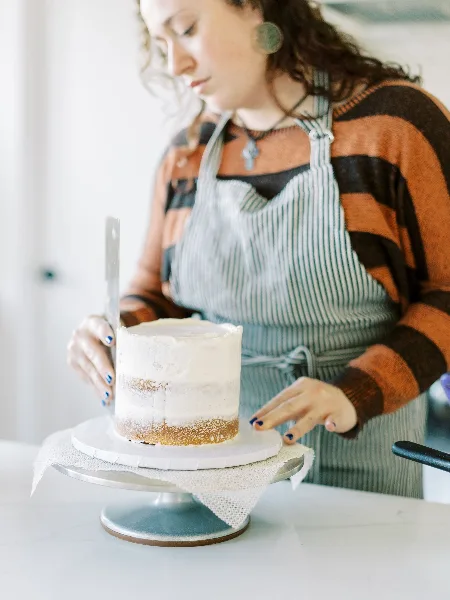 A baker wearing a striped apron is frosting a cake on a turntable.