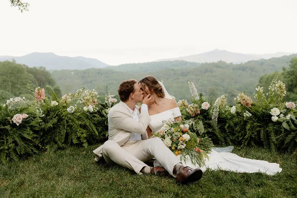 A bride and groom sitting on the grass in front of a floral arch with a mountain view in the background.