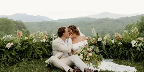 A bride and groom sitting on the grass in front of a floral arch with a mountain view in the background.