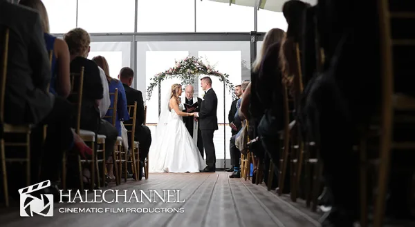 A bride and groom exchanging vows during a wedding ceremony, viewed from the aisle with guests seated on either side.