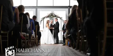 A bride and groom exchanging vows during a wedding ceremony, viewed from the aisle with guests seated on either side.