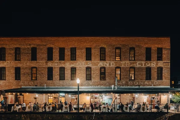 Nighttime exterior of a brick restaurant building with an outdoor patio where guests are seated at tables under string lights.