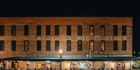Nighttime exterior of a brick restaurant building with an outdoor patio where guests are seated at tables under string lights.