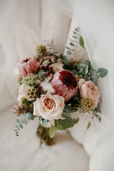 A close-up of a wedding bouquet with pink proteas, cream roses, and greenery resting on a white surface.