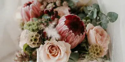 A close-up of a wedding bouquet with pink proteas, cream roses, and greenery resting on a white surface.