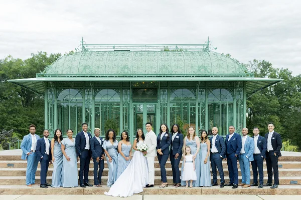 A wedding party consisting of the bride, groom, and their attendants posing for a group photo in front of a glass conservatory structure.