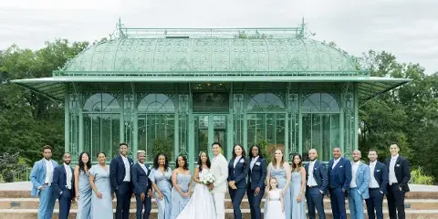 A wedding party consisting of the bride, groom, and their attendants posing for a group photo in front of a glass conservatory structure.