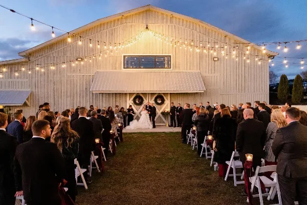 A wedding ceremony in progress with a bride and groom at the altar in front of a white barn venue decorated with string lights.