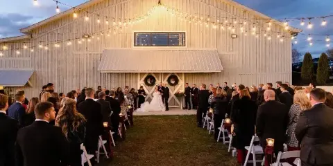 A wedding ceremony in progress with a bride and groom at the altar in front of a white barn venue decorated with string lights.