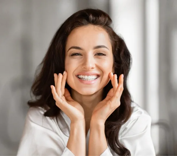 A smiling woman with styled hair and makeup touching her face.