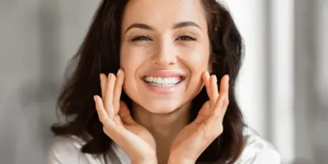 A smiling woman with styled hair and makeup touching her face.