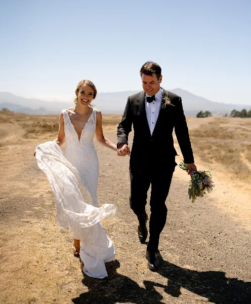 A bride and groom holding hands and walking down a dirt path in a sunny, open field.