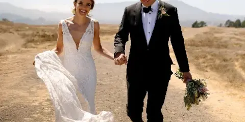 A bride and groom holding hands and walking down a dirt path in a sunny, open field.