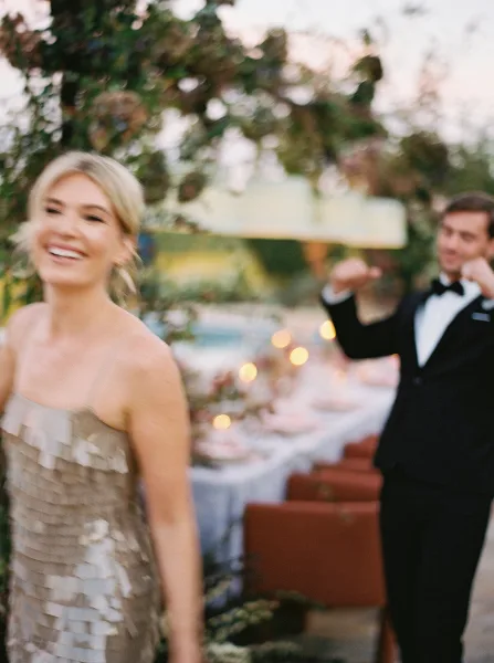 A smiling bride with a soft, natural updo and makeup, standing in front of a wedding reception table.
