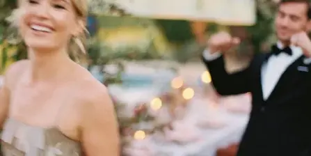 A smiling bride with a soft, natural updo and makeup, standing in front of a wedding reception table.