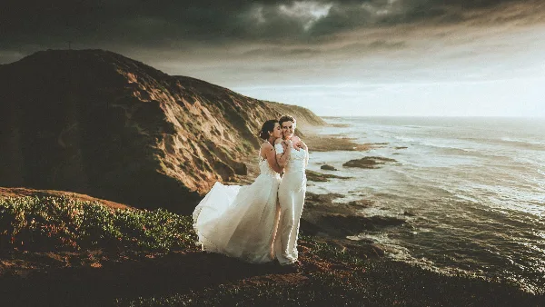A wedding couple embracing on a coastal cliff overlooking the ocean under a dramatic sky.