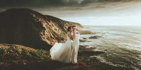 A wedding couple embracing on a coastal cliff overlooking the ocean under a dramatic sky.