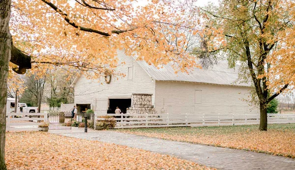 Exterior view of a white historic barn with a stone chimney, surrounded by white fencing and autumn trees.